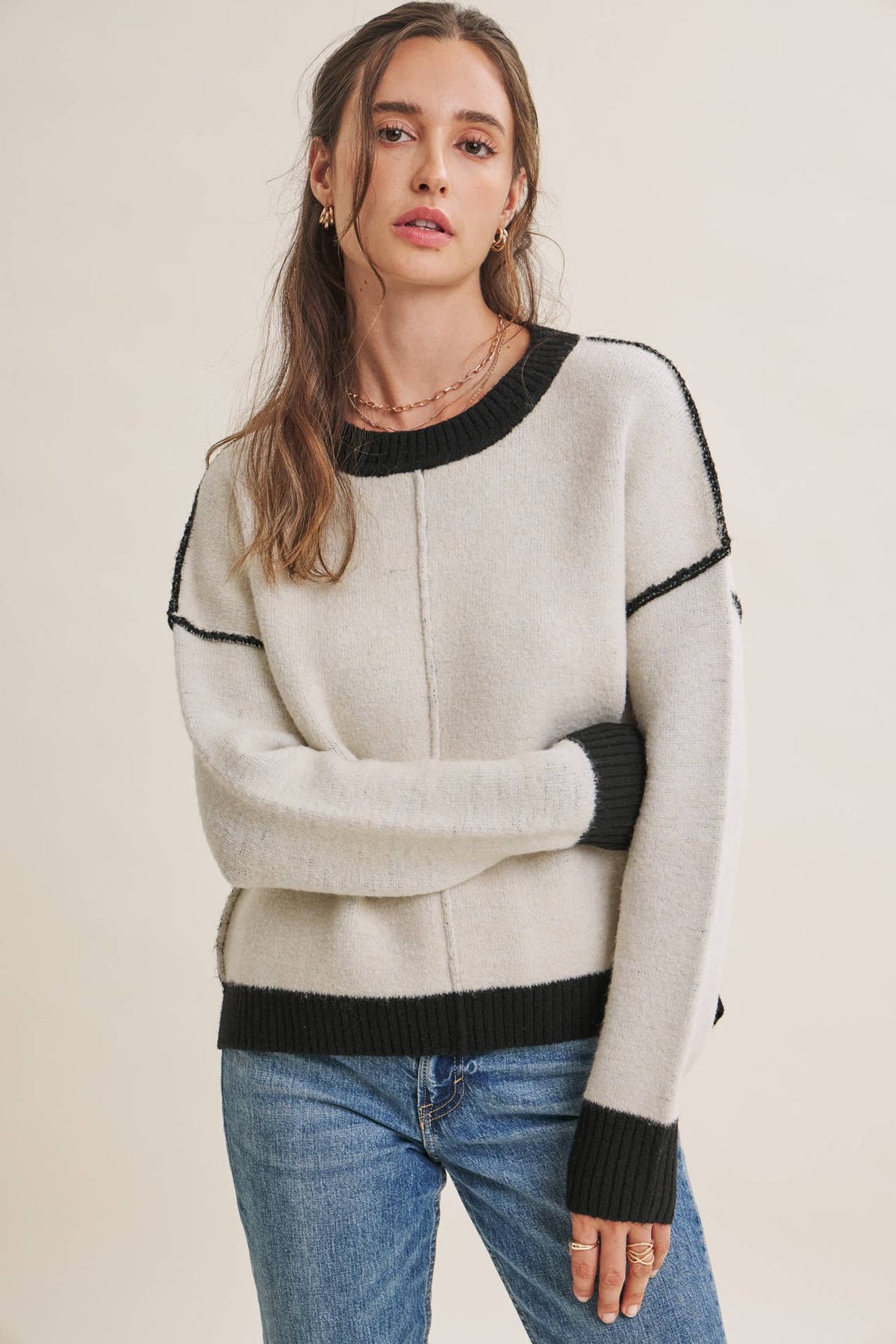A woman wearing a light beige knit sweater with black ribbed trim at the crew neck, cuffs, hem, and shoulder seams, paired with blue jeans, against a plain beige background at the rustic barn in killingworth ct