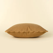 A square tan linen throw pillow with a scalloped edge, photographed against a solid cream background at the rustic barn in killingworth ct