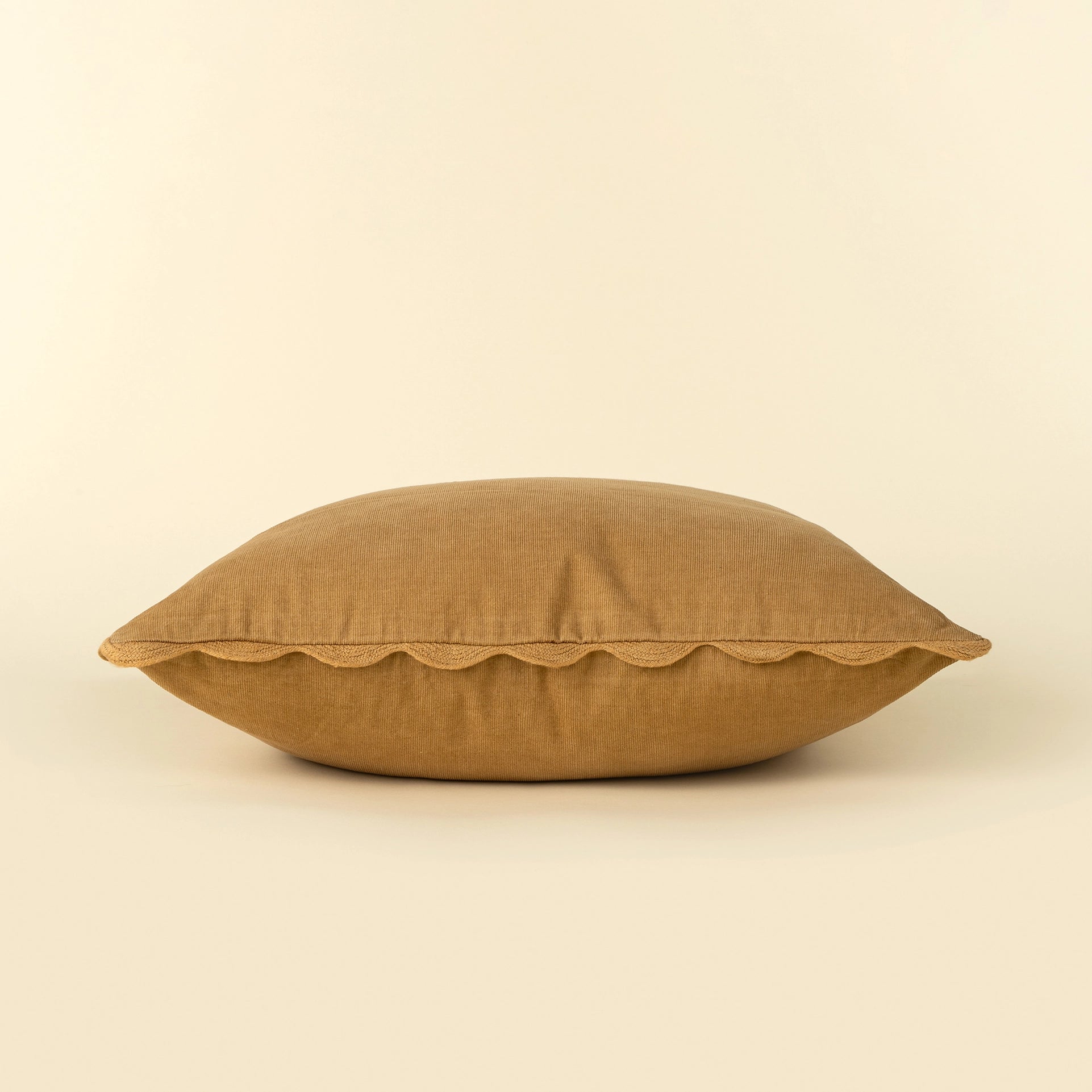 A square tan linen throw pillow with a scalloped edge, photographed against a solid cream background at the rustic barn in killingworth ct