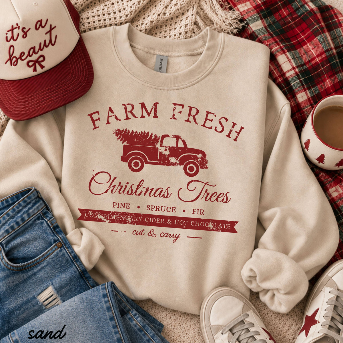 A cream-colored crewneck sweatshirt with a red graphic print reading 'FARM FRESH Christmas Trees' and listing holiday ingredients, styled in a cozy flat-lay with jeans, a red trucker hat, a mug, and white sneakers.