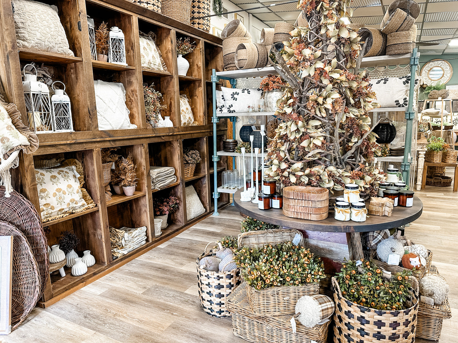 A styled home decor store interior featuring a rustic wooden shelving unit filled with pillows, lanterns, and textiles, alongside a round display table with a decorative tree and wicker baskets on a light wood floor.