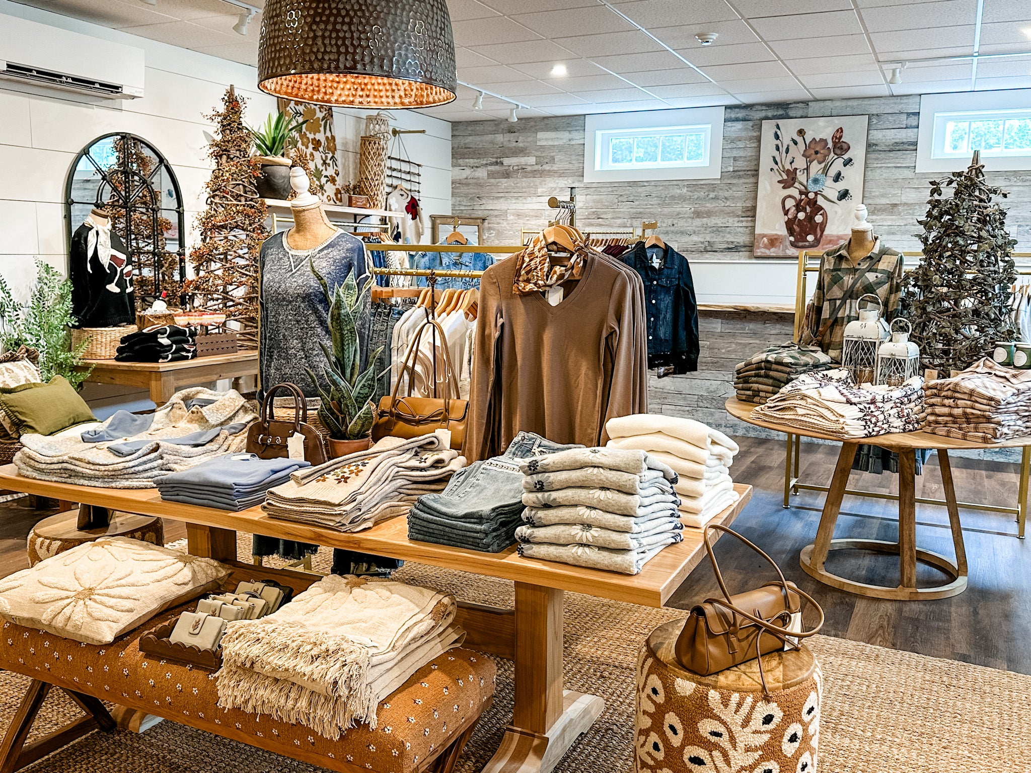A cozy boutique interior displaying folded knit sweaters, blankets, scarves, and handbags on wooden tables and mannequins. The shop features warm lighting, a dark woven pendant lamp, a wood-paneled wall, and decorative Christmas trees.