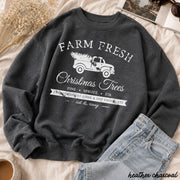 A dark heather charcoal crewneck sweatshirt with a white graphic print of a vintage truck carrying Christmas trees, styled in a flat lay with light blue jeans, dried flowers, and a cup of coffee.