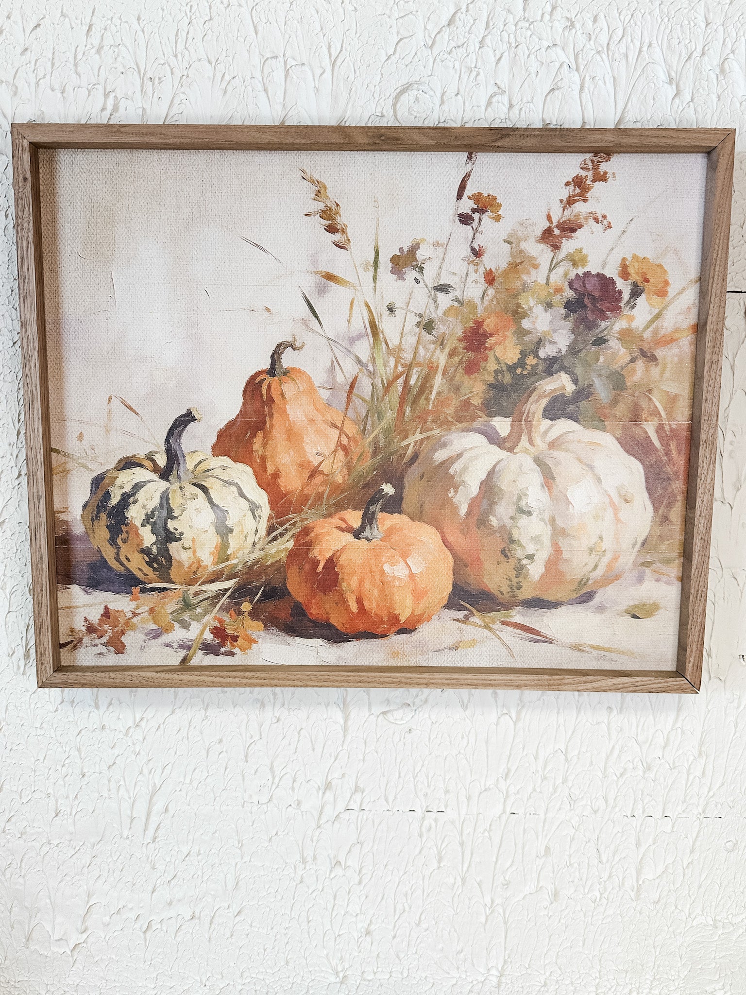 A framed still life painting featuring orange and white pumpkins with dried flowers and grasses, displayed on a textured white wall at the rustic barn in killingworth ct