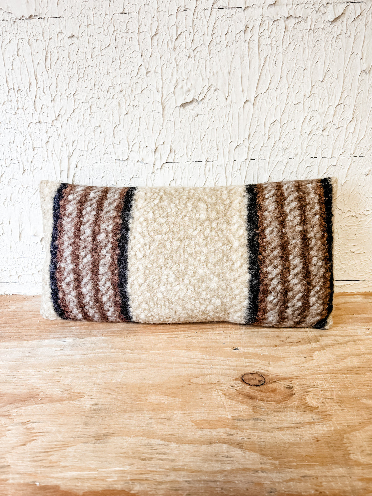 A rectangular woven throw pillow with a cream center and brown striped sides, resting on a light wood surface against a textured white wall. At the rustic barn in killingworth ct