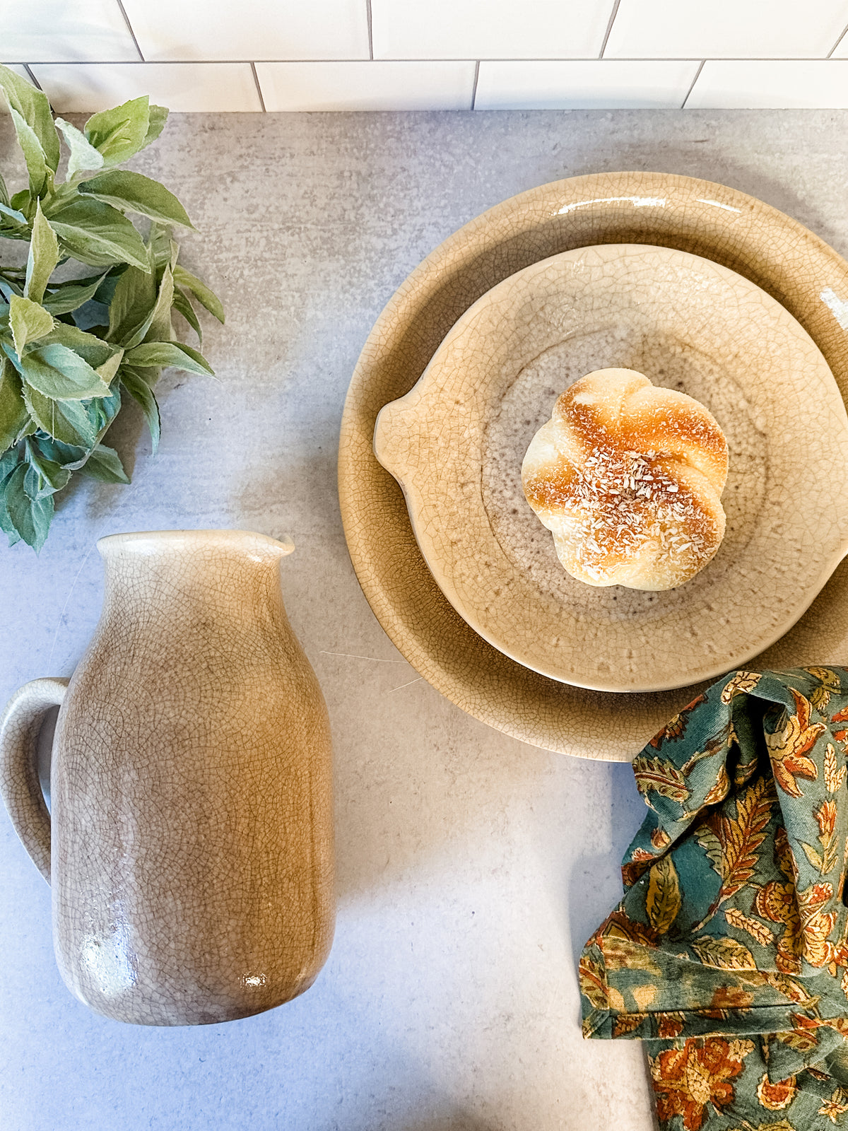 A styled flat-lay featuring a ceramic pitcher and a stack of ceramic plates with a small bread roll on top, set on a light gray countertop with a green cloth and basil leaves at the rustic barn in killingworth, CT