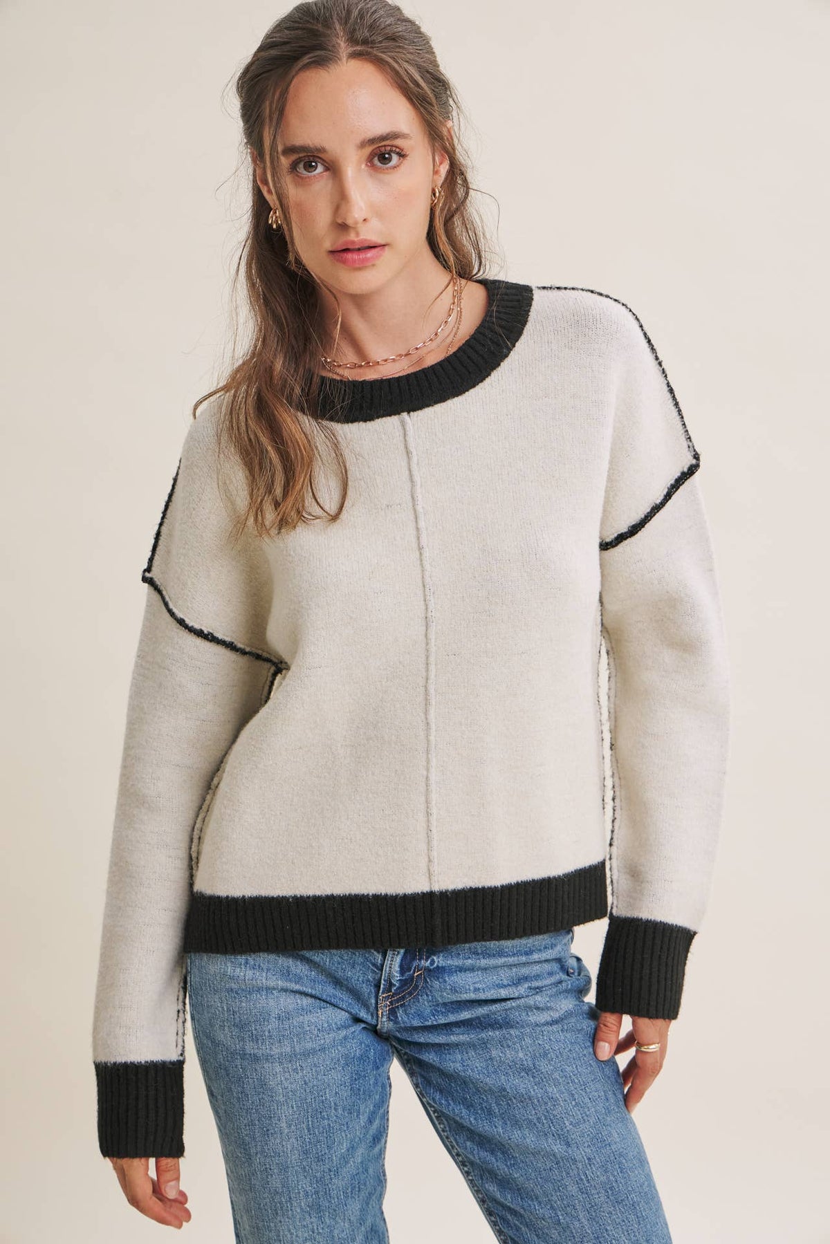 A woman wearing a cream-colored knit sweater with black ribbed trim and blue jeans, standing against a plain light beige background at the rustic barn in killingworth ct