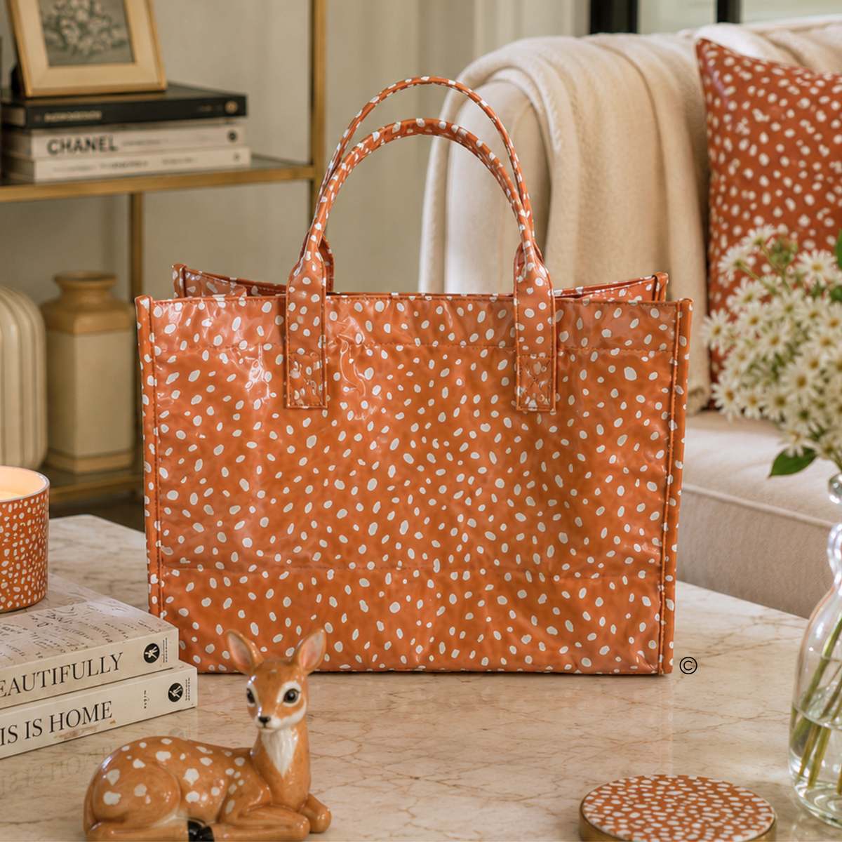 An orange tote bag with a white dalmatian spot pattern sits on a marble table in a cozy living room setting, styled with a deer figurine, books, and a sofa in the background.