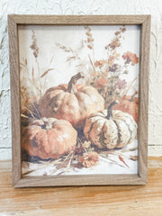 A framed autumn still life print featuring three pumpkins in warm peach, orange, and cream tones with dried flowers and grasses, set in a rustic light brown wood frame on a wooden surface against a textured white wall. At the rustic barn in killingworth ct