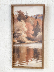 A framed vertical landscape painting of an autumn lakeside scene with peach, orange, and brown trees reflected in calm water, mounted on a textured white wall at the rustic barn in killingworth ct