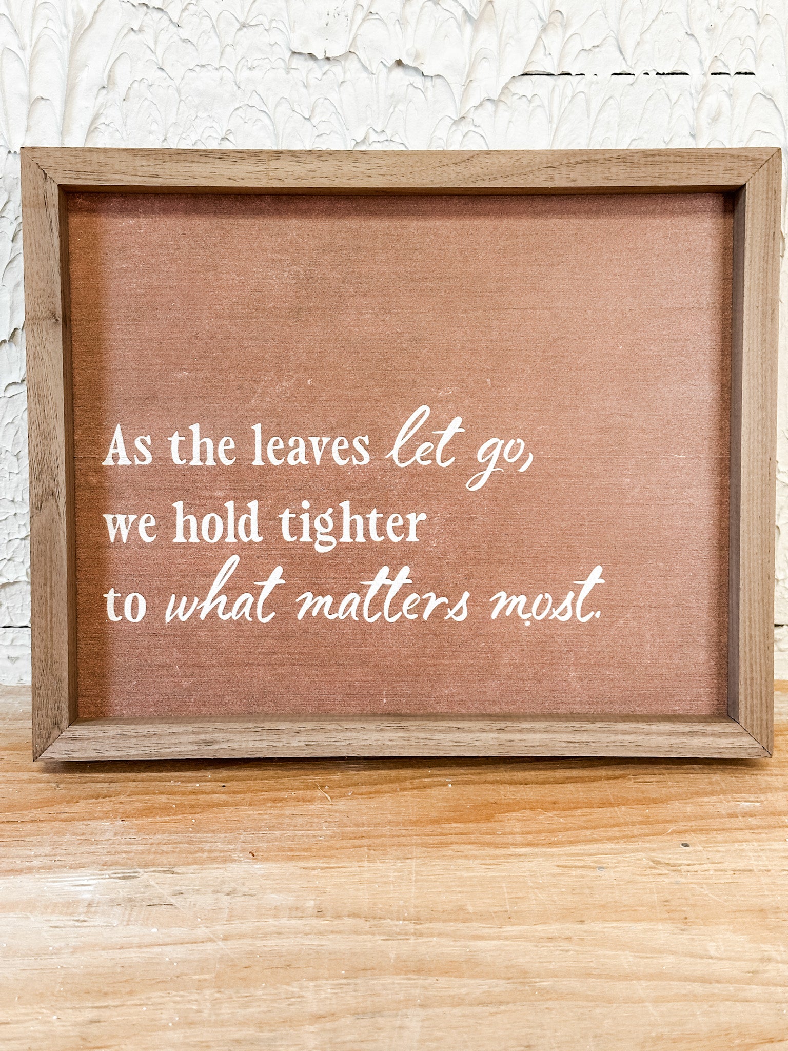 A square framed rustic sign with a brown linen-like center and white script text reading, 'As the leaves let go, we hold tighter to what matters most.' The sign has a natural wood frame and is photographed against a white textured wall and light wood surface.