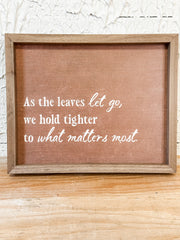 A square framed rustic sign with a brown linen-like center and white script text reading, 'As the leaves let go, we hold tighter to what matters most.' The sign has a natural wood frame and is photographed against a white textured wall and light wood surface.