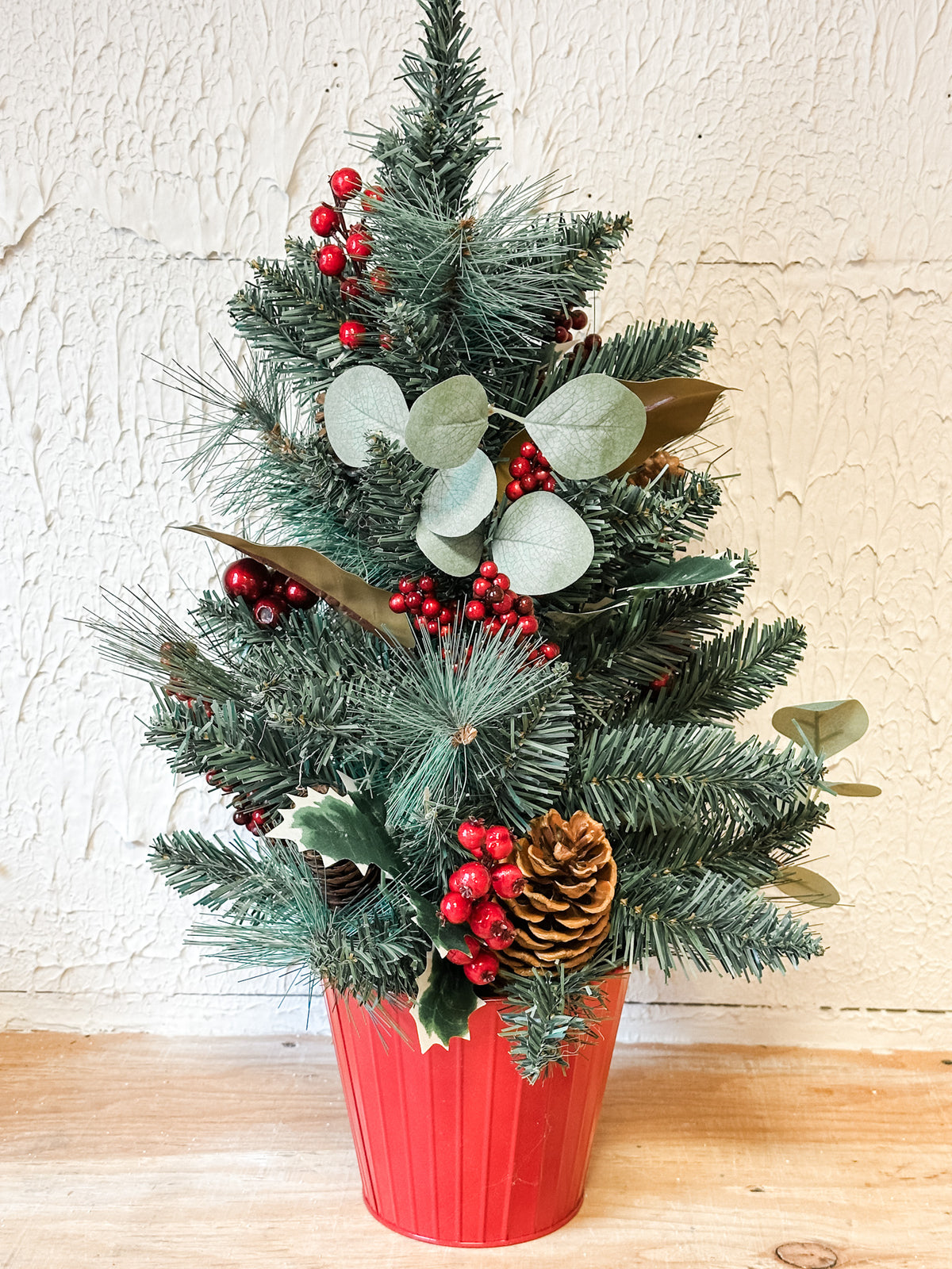 A small artificial Christmas tree arrangement in a red ribbed pot, decorated with green pine branches, silver eucalyptus leaves, red berries, holly leaves, and a pinecone, set against a textured off-white wall and light wood surface. Available at the rustic barn in killingworth ct