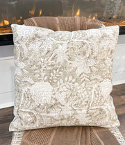 A square decorative throw pillow with an intricate floral and botanical damask pattern in beige and cream, resting on a woven rattan chair in front of a lit fireplace from the rustic barn in killingworth,CT