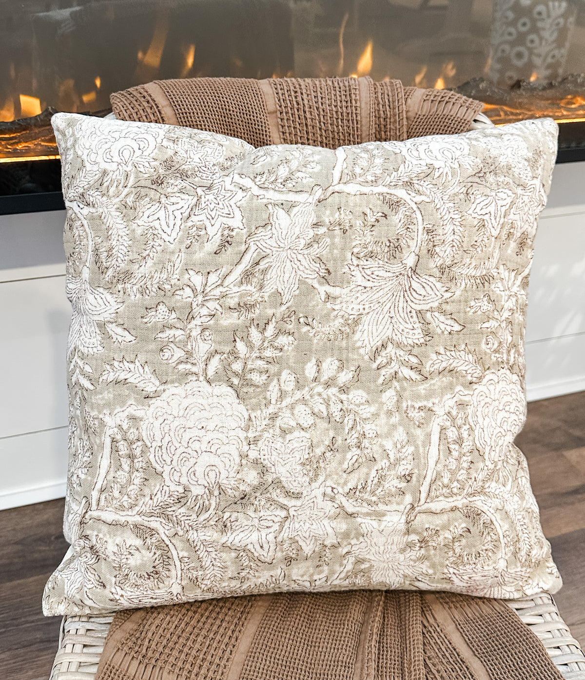 A square decorative throw pillow with an intricate floral and botanical damask pattern in beige and cream, resting on a woven rattan chair in front of a lit fireplace from the rustic barn in killingworth,CT