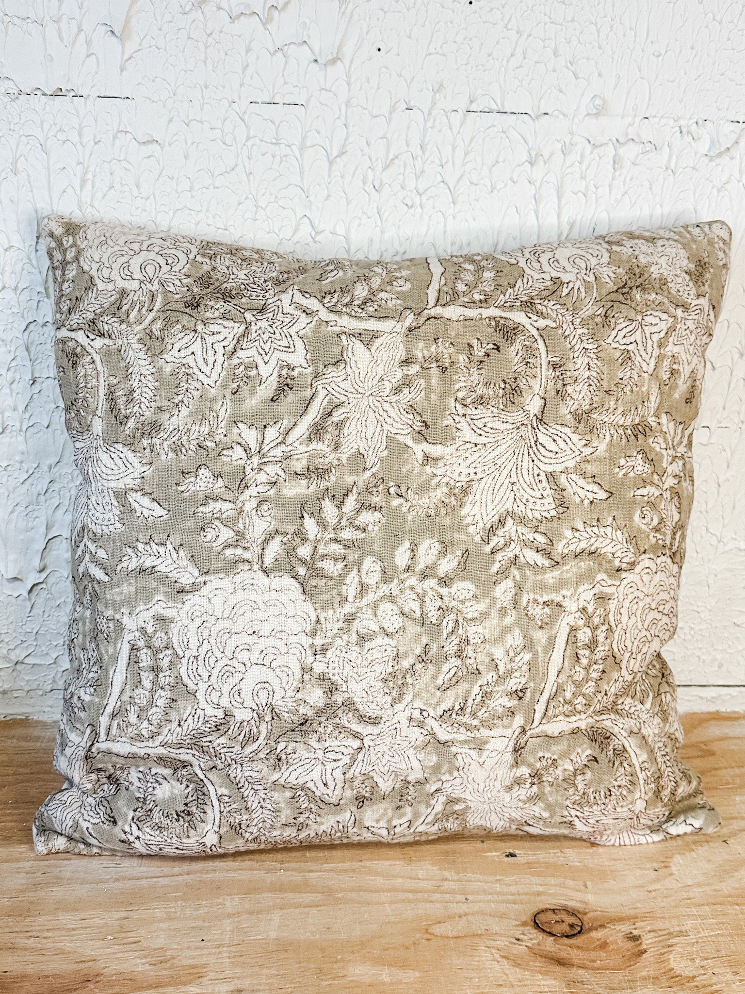 A square decorative throw pillow with an intricate floral and botanical damask pattern in beige and cream, resting on a woven rattan chair in front of a lit fireplace from the rustic barn in killingworth, CT