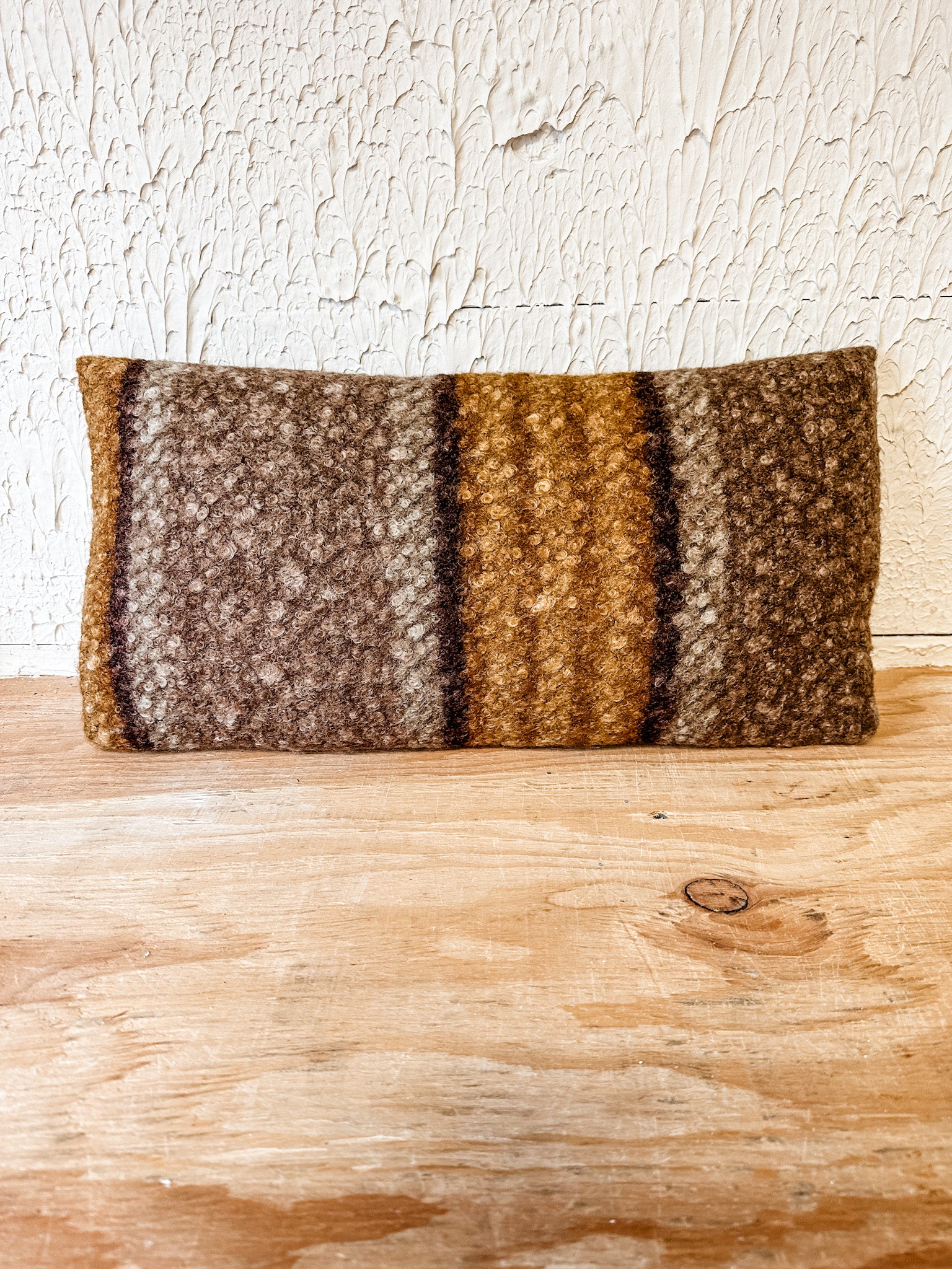 A rectangular woven throw pillow with a rustic striped pattern in earthy brown, beige, and golden tones, resting on a light wooden surface against a textured off-white wall. At the rustic barn in killingworth ct