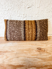 A rectangular woven throw pillow with a rustic striped pattern in earthy brown, beige, and golden tones, resting on a light wooden surface against a textured off-white wall. At the rustic barn in killingworth ct
