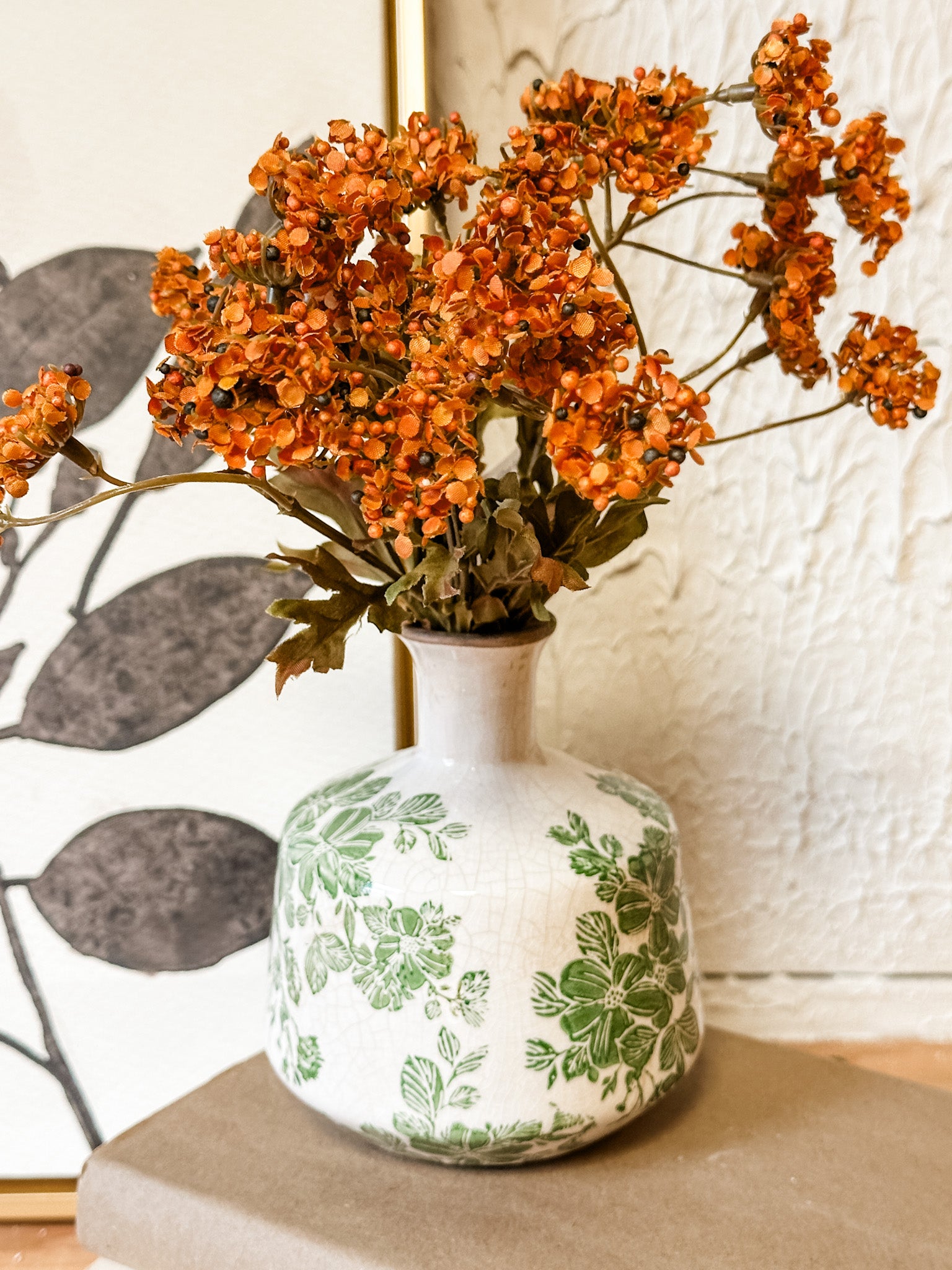 A white ceramic vase with a green floral toile pattern, filled with dried orange flowers, set on a neutral surface against a textured white wall at the rustic barn in killingworth ct