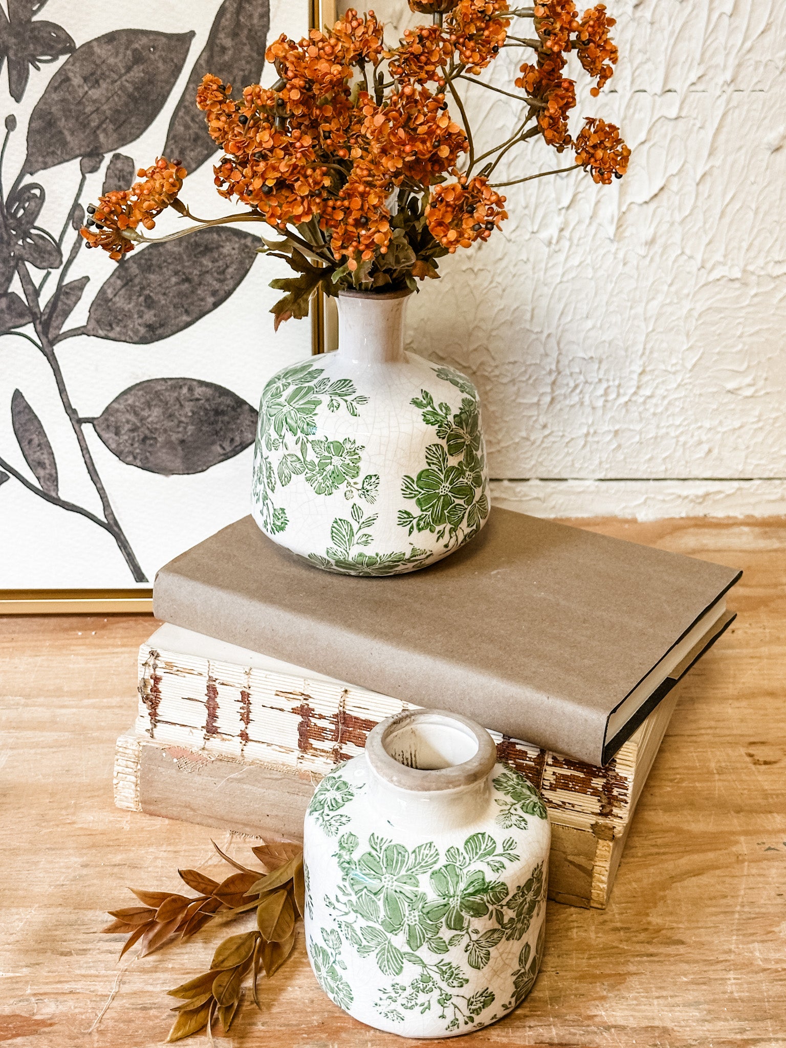 Two white ceramic vases with green botanical patterns, styled on a wooden surface with books and dried orange flowers against a textured white wall at the rustic barn in killingworth ct