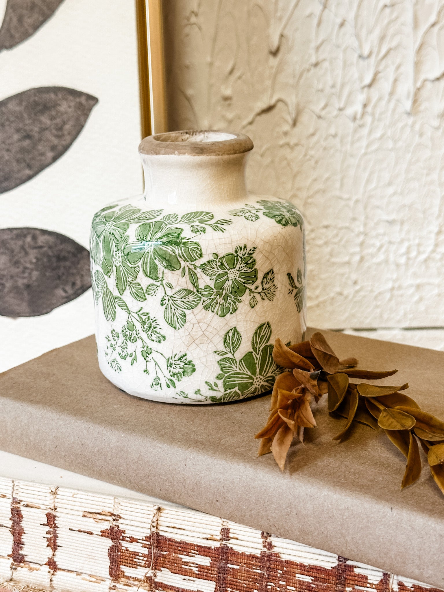 A small ceramic vase with a green botanical floral pattern on a cream background, styled on a brown box with dried leaves in a home decor setting.\ at the rustic barn in killingworth ct