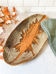An oval ceramic dish with a textured, porous surface in earthy brown and beige tones, featuring a raised orange-brown sunburst design in the center. The dish is styled on a light gray surface with a sage green towel and autumn leaves. At the rustic barn in killingworth ct