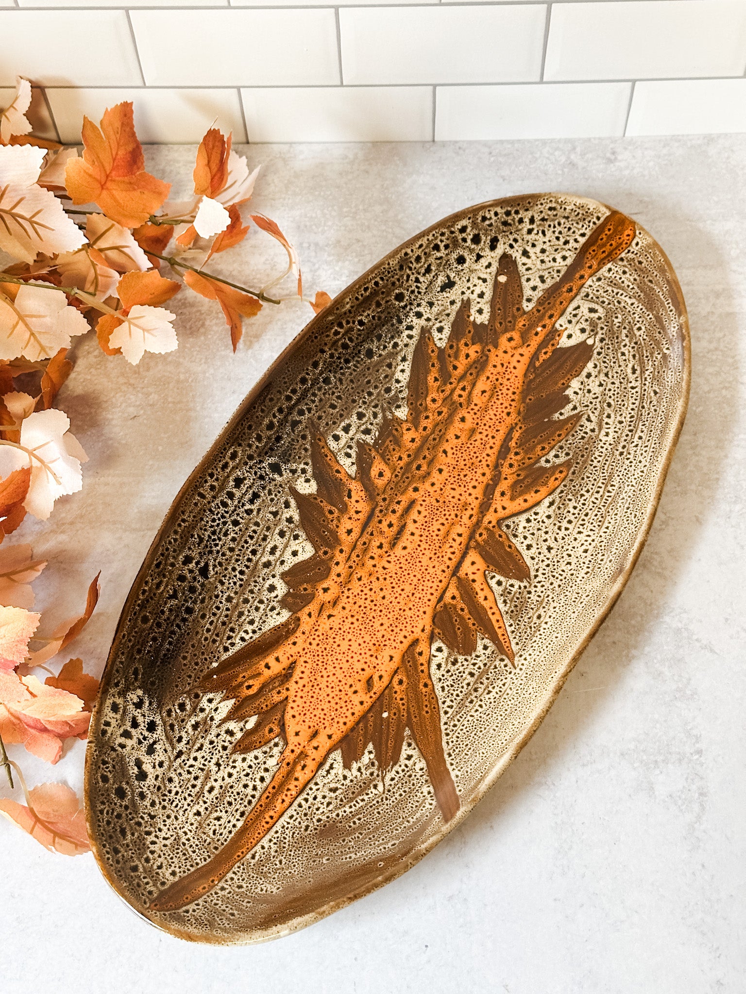 An oval ceramic dish with a textured, porous surface in earthy brown and beige tones, featuring a raised orange-brown sunburst design in the center. The dish is styled on a light gray surface with a sage green towel and autumn leaves. At the rustic barn in killingworth ct