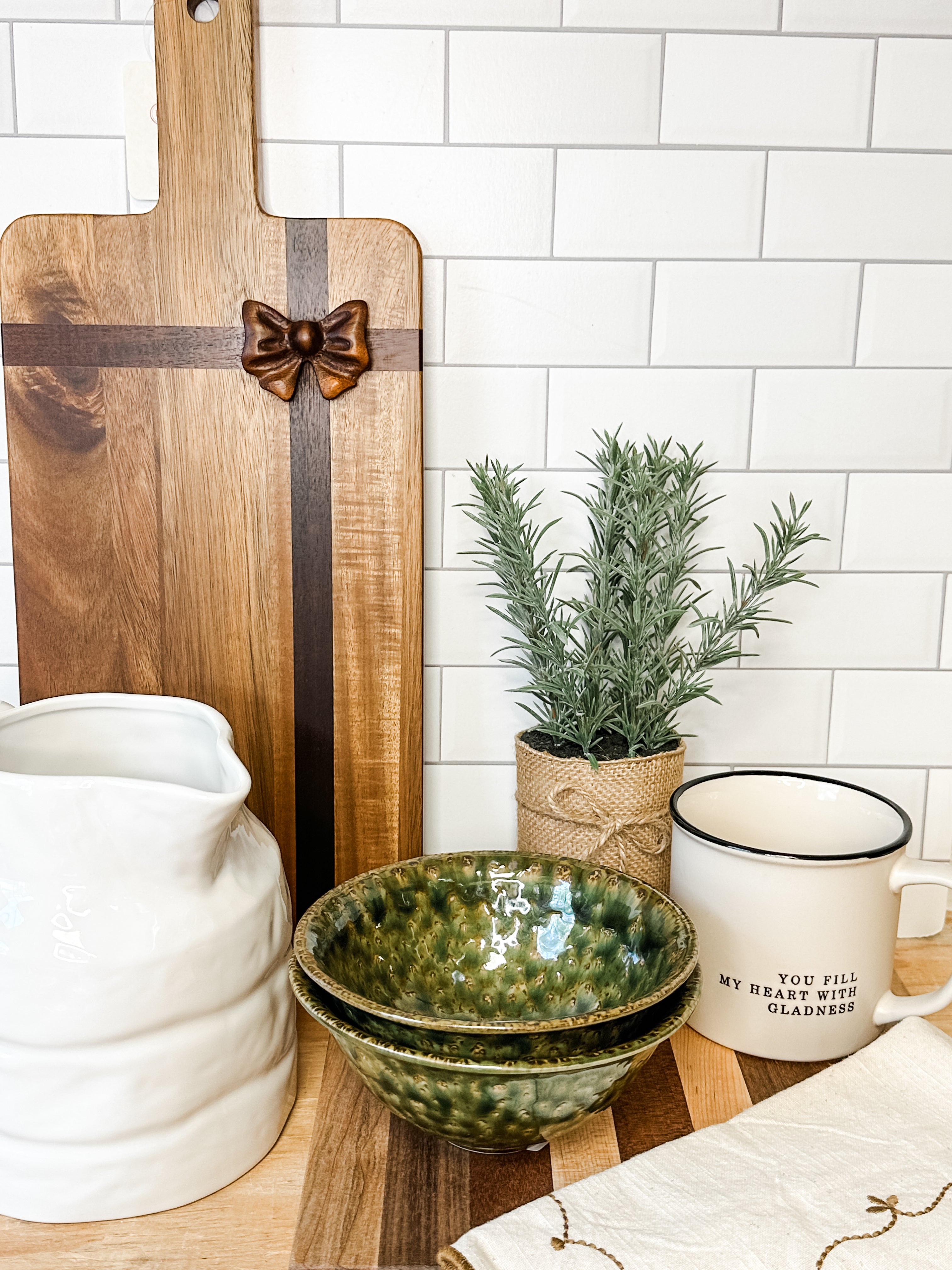 A styled kitchen countertop scene featuring a green glazed ceramic bowl set, a white ceramic pitcher, a white enamel mug with black trim and text, a wooden cutting board with a bow inlay, a small potted rosemary plant, and a white subway tile backsplash behind at the rustic barn in killingworth ct