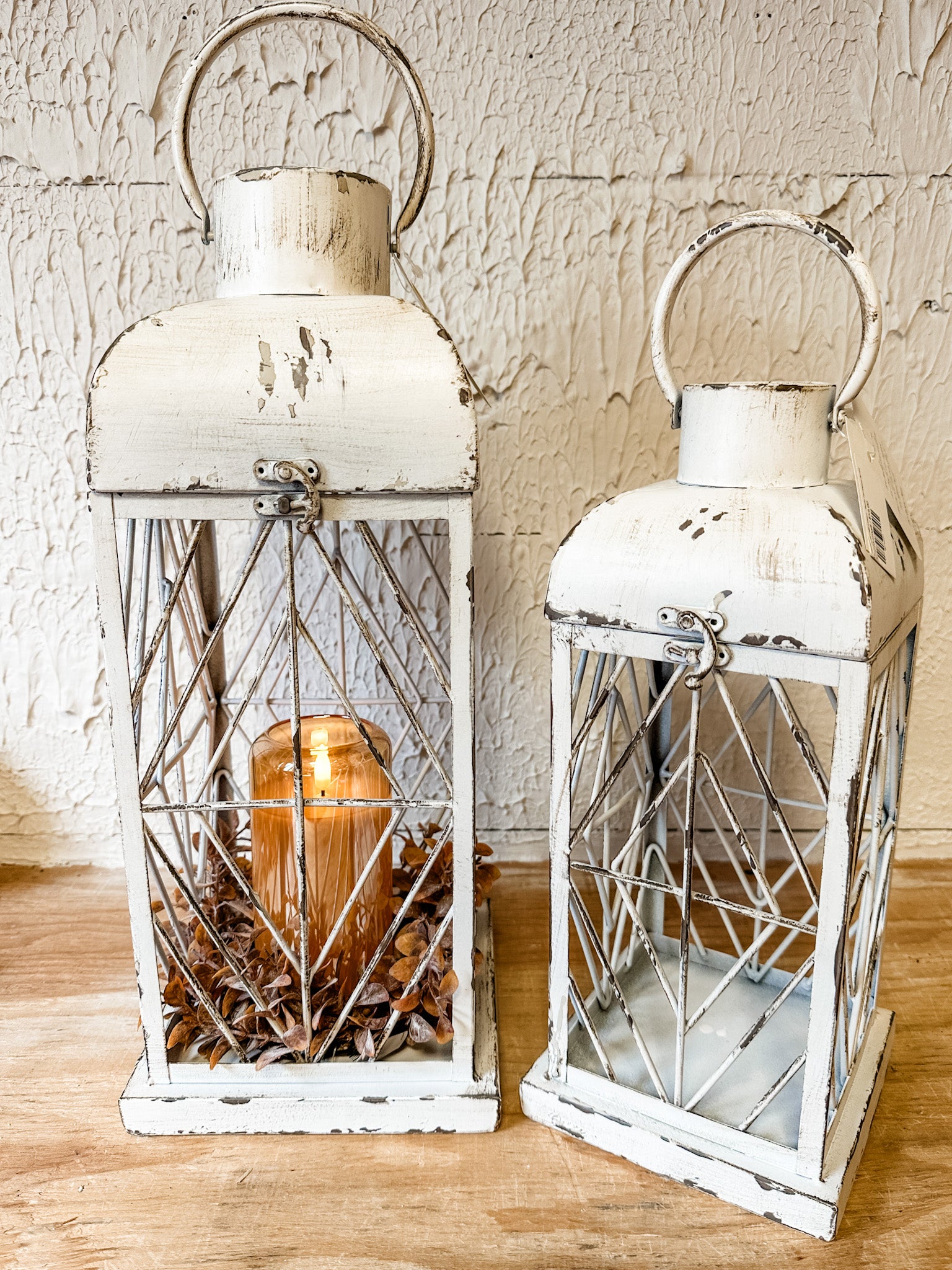Two distressed white metal lanterns with wire crisscross fronts and arched handles, displayed on a wooden surface against a textured off-white wall. One lantern is empty, while the other contains a lit amber glass candle.  At the rustic barn in killingworth ct