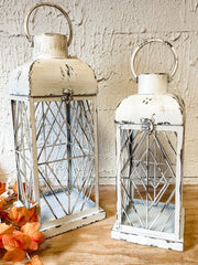 Two distressed white metal lanterns with wire crisscross patterns and top handles, displayed on a wooden surface against a textured beige wall, with orange autumn leaves in the lower left corner at the rustic barn in killingworth ct