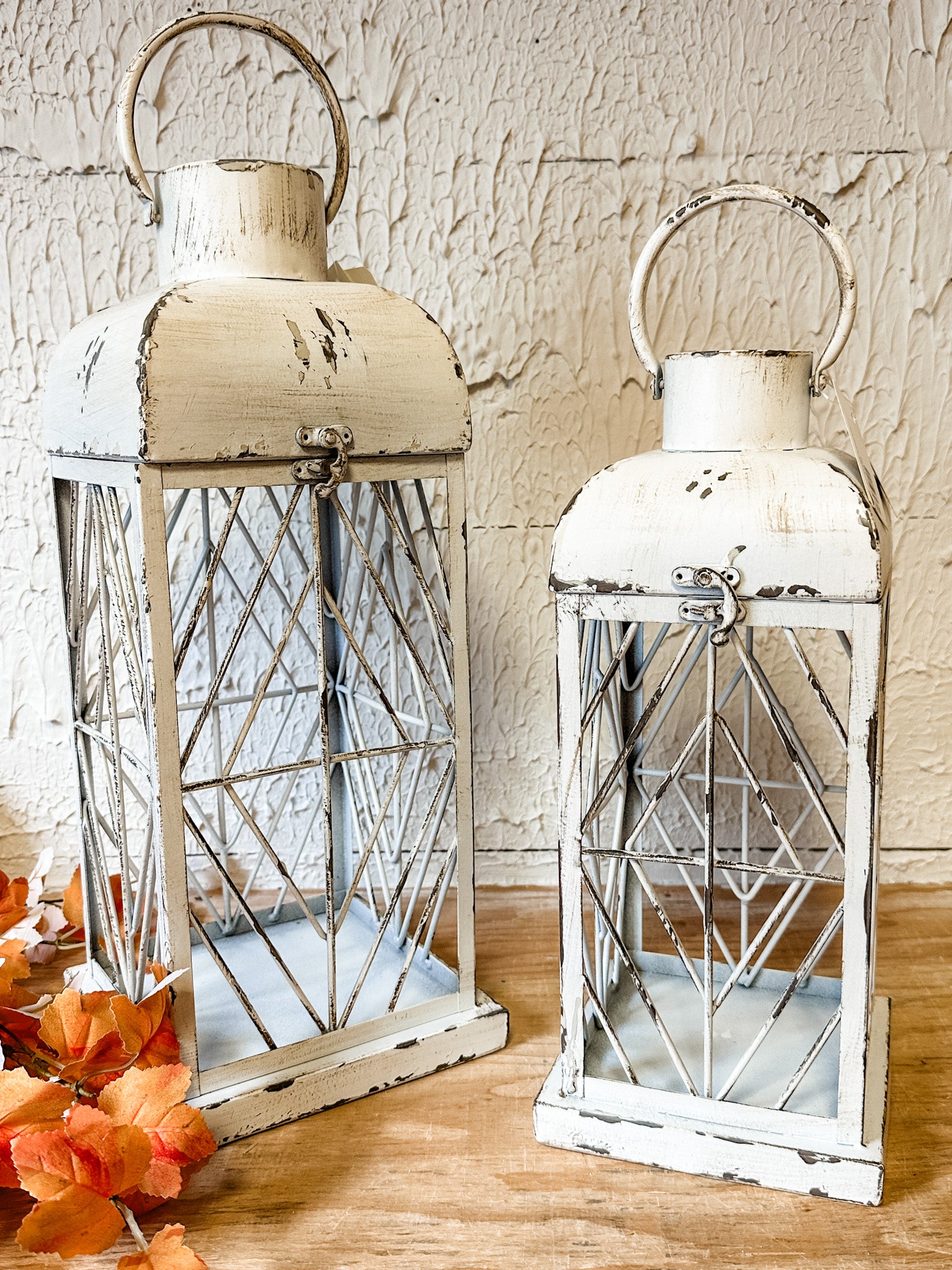 Two distressed white metal lanterns with wire crisscross patterns and top handles, displayed on a wooden surface against a textured beige wall, with orange autumn leaves in the lower left corner at the rustic barn in killingworth ct
