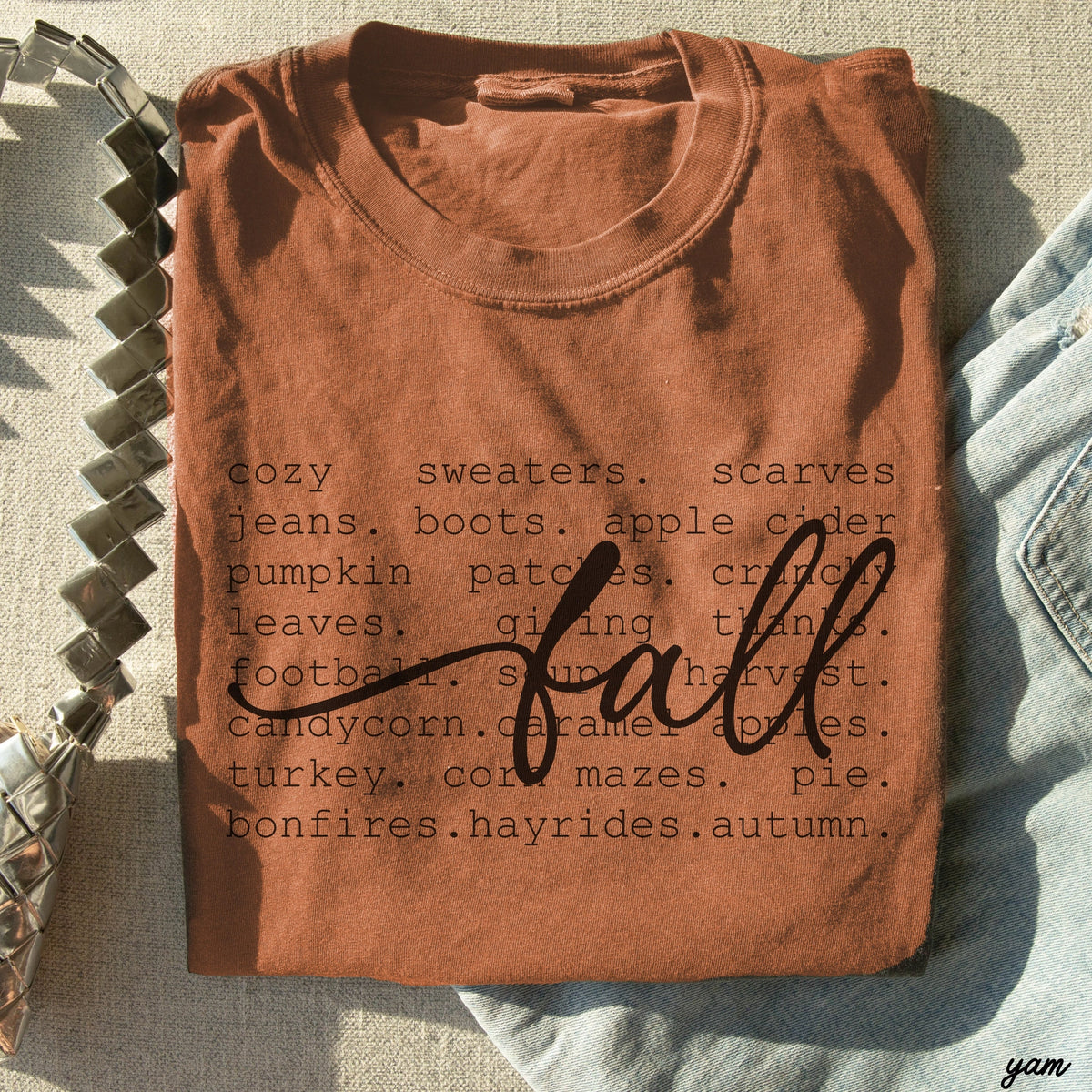 A folded terracotta brown t-shirt with a dark graphic print reading 'cozy sweaters. scarves jeans at the rustic barn in killingworth ct