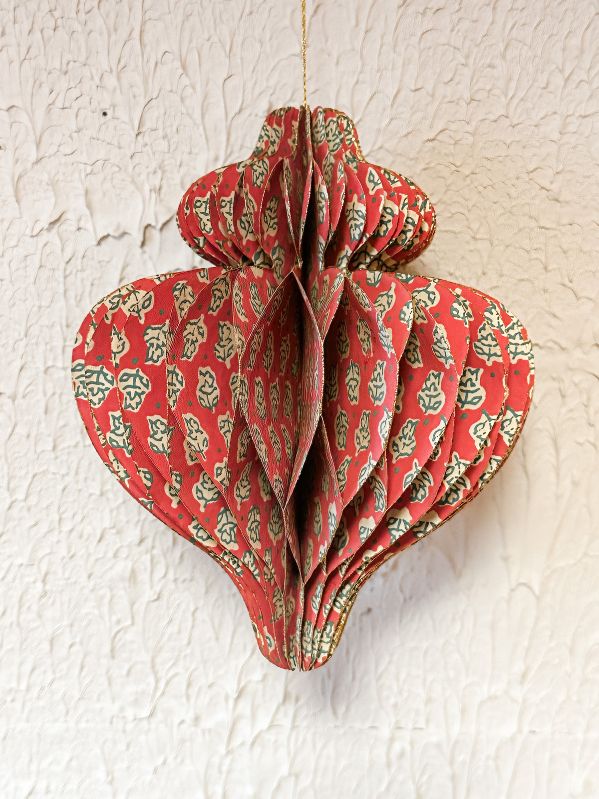 A red paper honeycomb hanging ornament decorated with a green and beige floral block print, suspended against a textured off-white wall at the rustic barn in killingworth ct