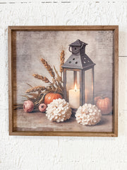 Framed rustic wall art featuring a still life scene with a black lantern, lit candle, wheat stalks, pumpkins, and white flowers on a textured beige background, shown in a wooden frame against a white wall at the rustic barn in killingworth ct