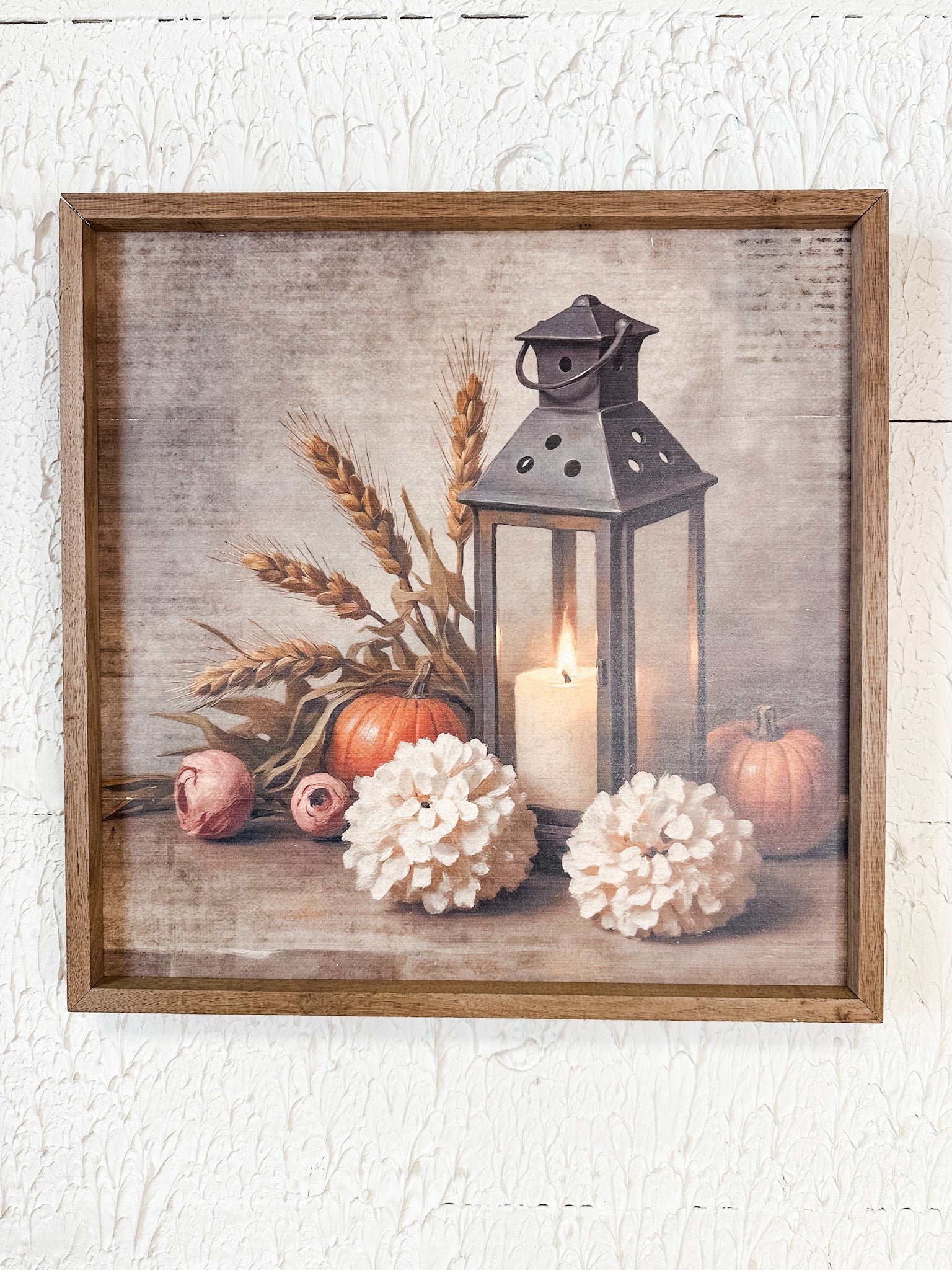 Framed rustic wall art featuring a still life scene with a black lantern, lit candle, wheat stalks, pumpkins, and white flowers on a textured beige background, shown in a wooden frame against a white wall at the rustic barn in killingworth ct