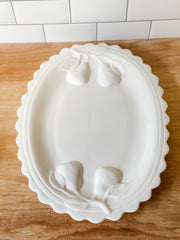 A white ceramic oval platter with a scalloped rim and raised pear motifs in the corners, shown on a wooden surface with a white subway tile backsplash behind it at the rustic barn in killingworth ct