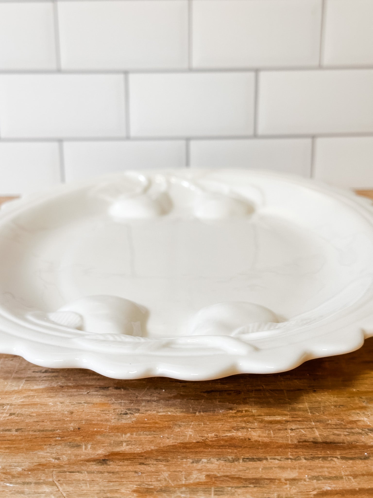 A white ceramic serving platter with embossed floral details and a scalloped rim, resting on a wooden surface in front of a white subway tile backsplash at the rustic barn in killingworth ct