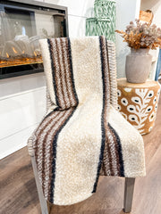 A cream-colored textured throw blanket with brown and black vertical stripes is draped over a chair in a cozy living room setting next to a fireplace and a patterned side table. At the rustic barn in killingworth ct