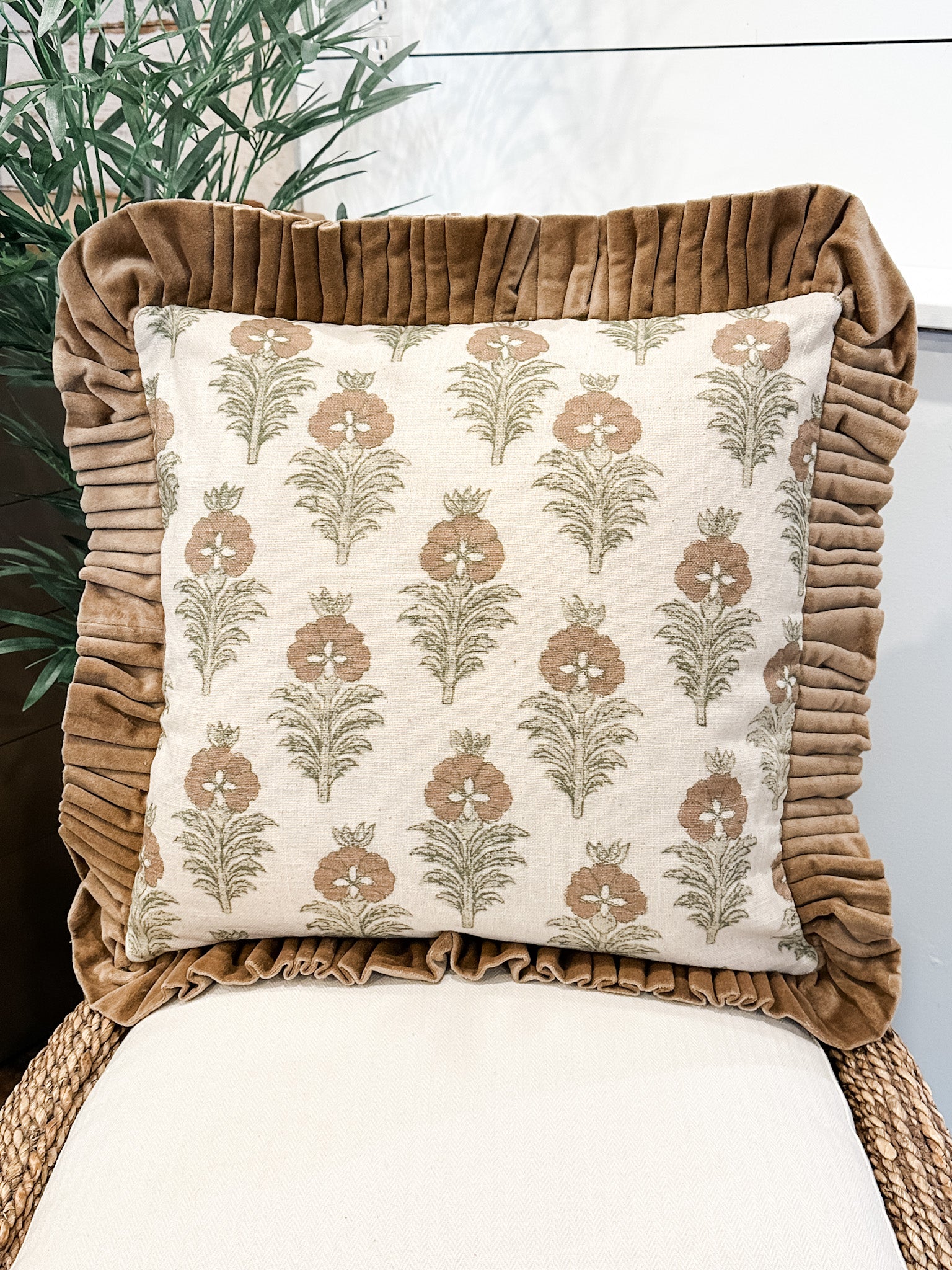 A square decorative throw pillow with a cream background and a brown botanical pattern, framed by a wide brown ruffled velvet border, displayed on a woven chair at the rustic barn in killingworth ct