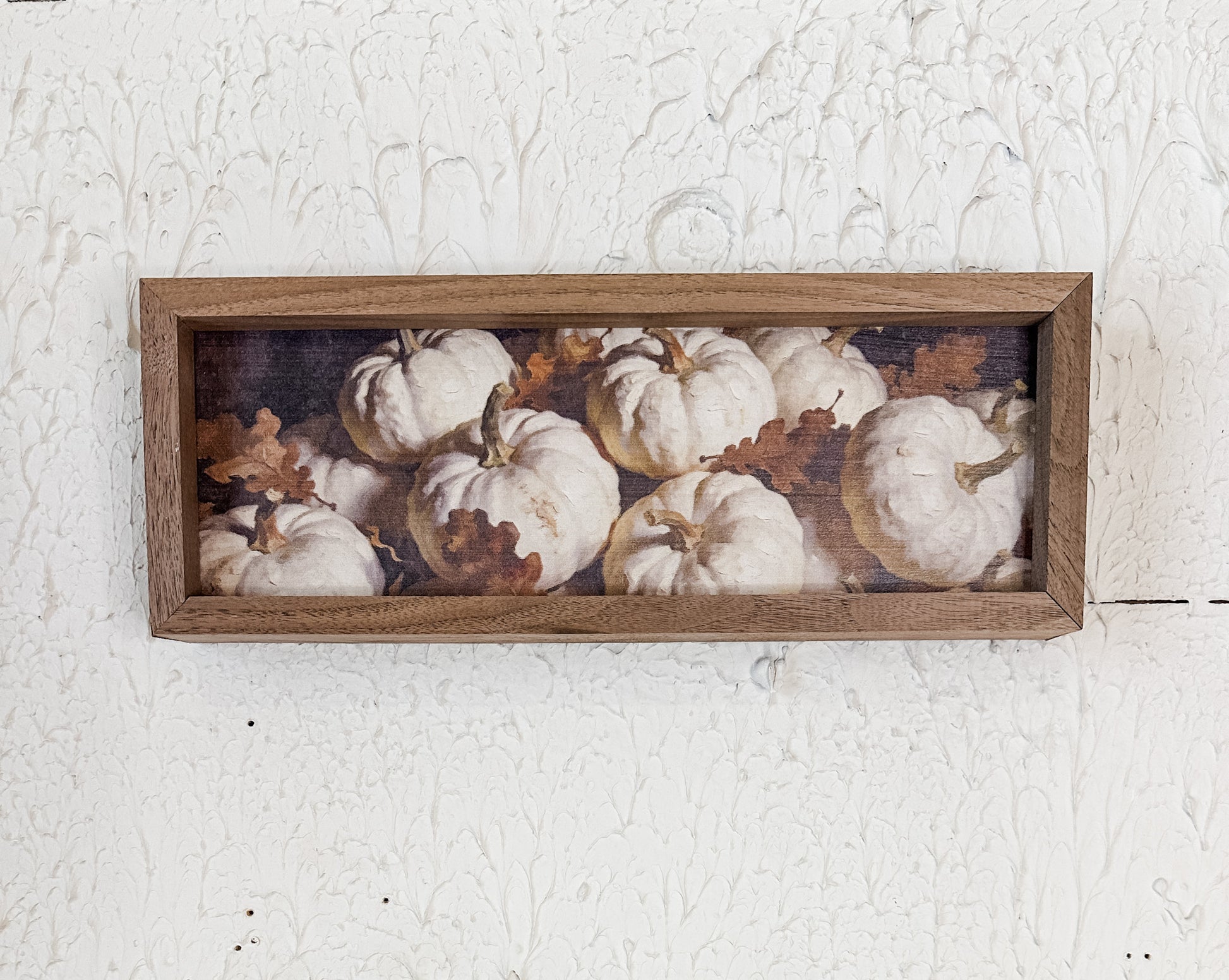 A rectangular wooden framed wall art piece featuring a printed still life of white pumpkins and brown leaves, mounted on a textured white wall.  At the rustic barn in Killingworth Ct