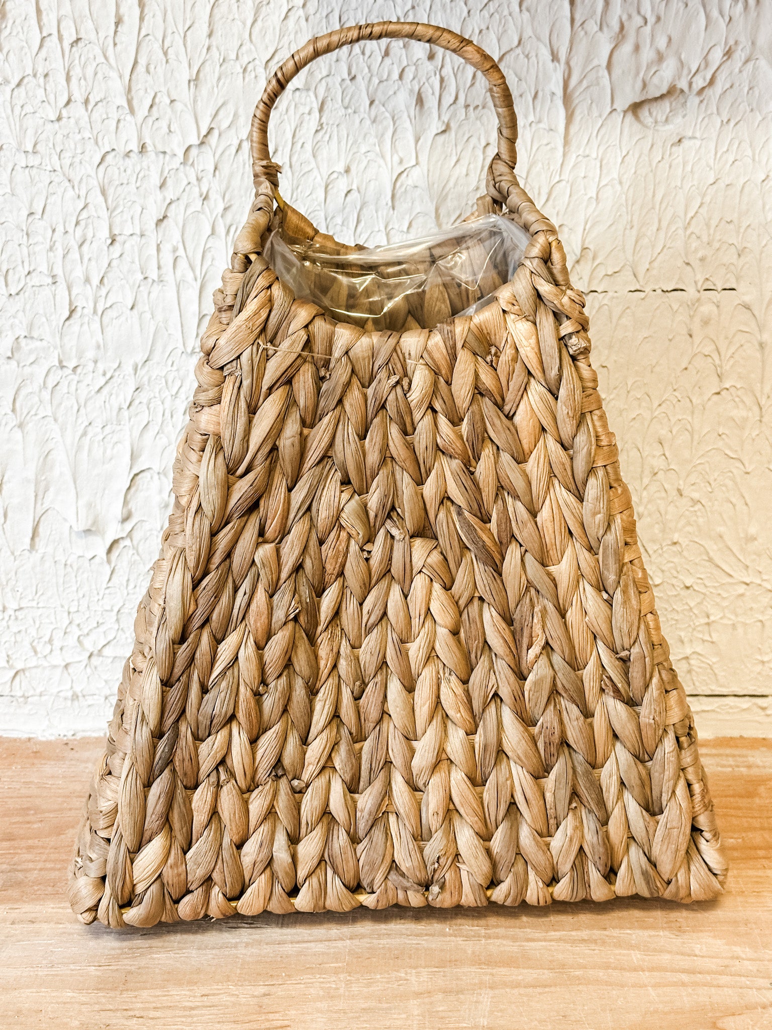 A natural woven straw handbag with a single top handle, resting on a light wood surface against a textured off-white wall at the rustic barn in killingworth ct