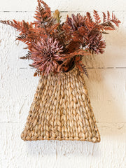 A natural woven seagrass basket or vase with a flared triangular base, filled with dried autumn foliage in rust, terracotta, and brown tones, set against a white textured wall at the rustic barn in killingworth ct