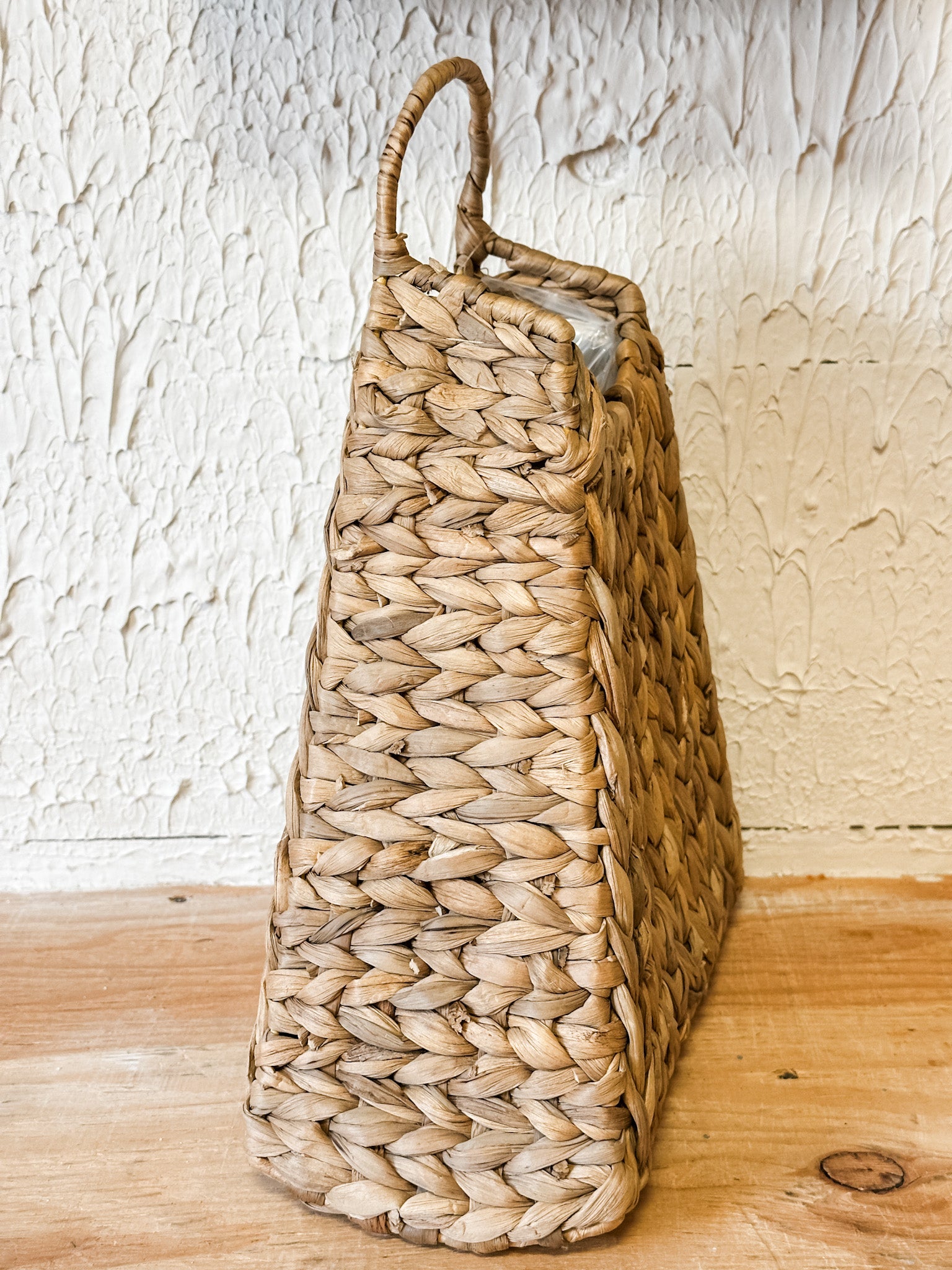 A tall, narrow woven basket made from natural straw or seagrass with a single loop handle, standing on a light wood surface against a textured off-white wall at the rustic barn in killingworth ct