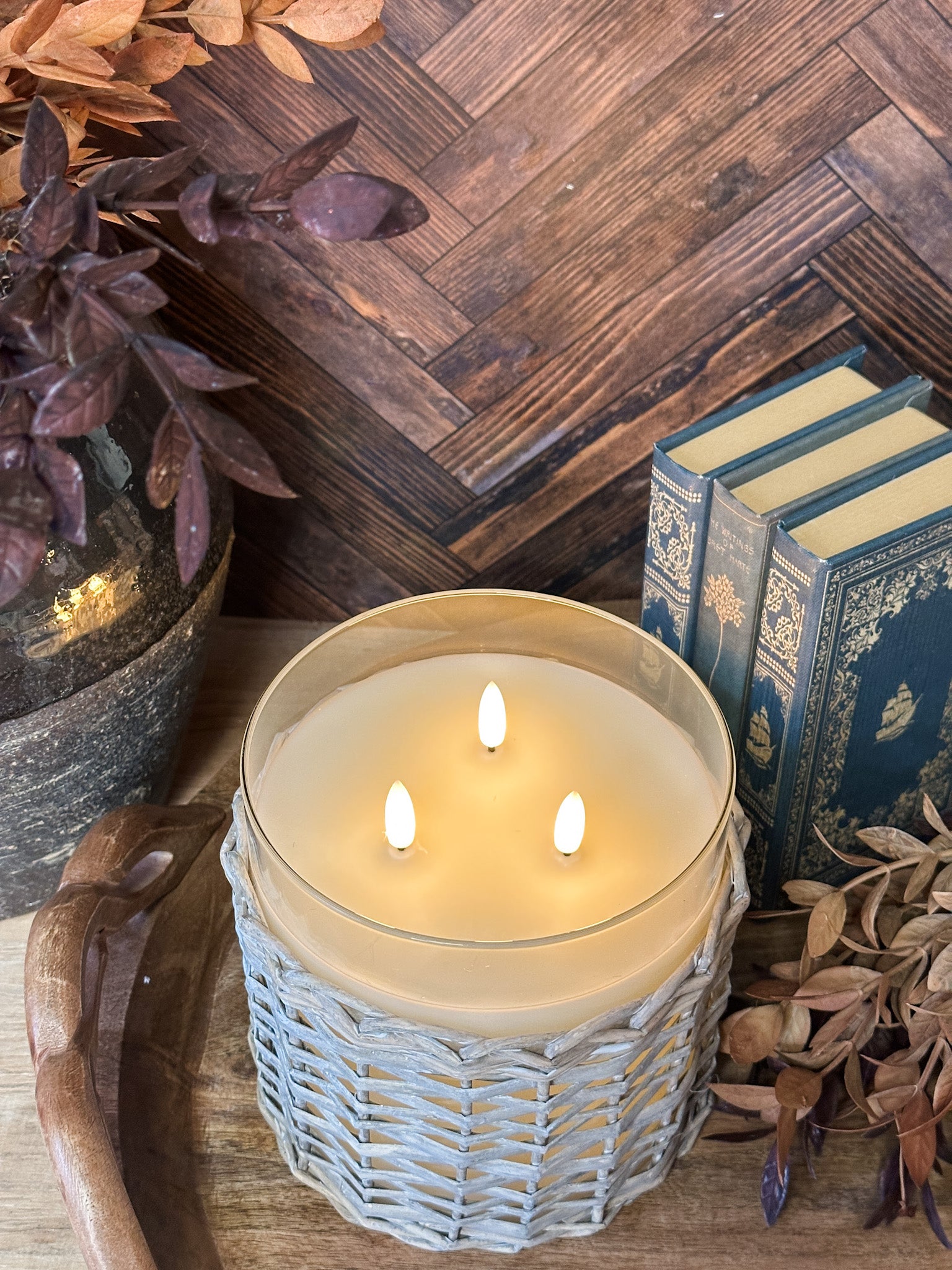 A lit candle in a woven light gray holder sits on a wooden surface surrounded by dried pinecones and branches, with a dark wood herringbone wall and books in the background at the rustic barn in killingworth, CT