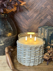 A lit candle in a woven light gray holder sits on a wooden surface surrounded by dried pinecones and branches, with a dark wood herringbone wall and books in the background at the rustic barn in killingworth, CT