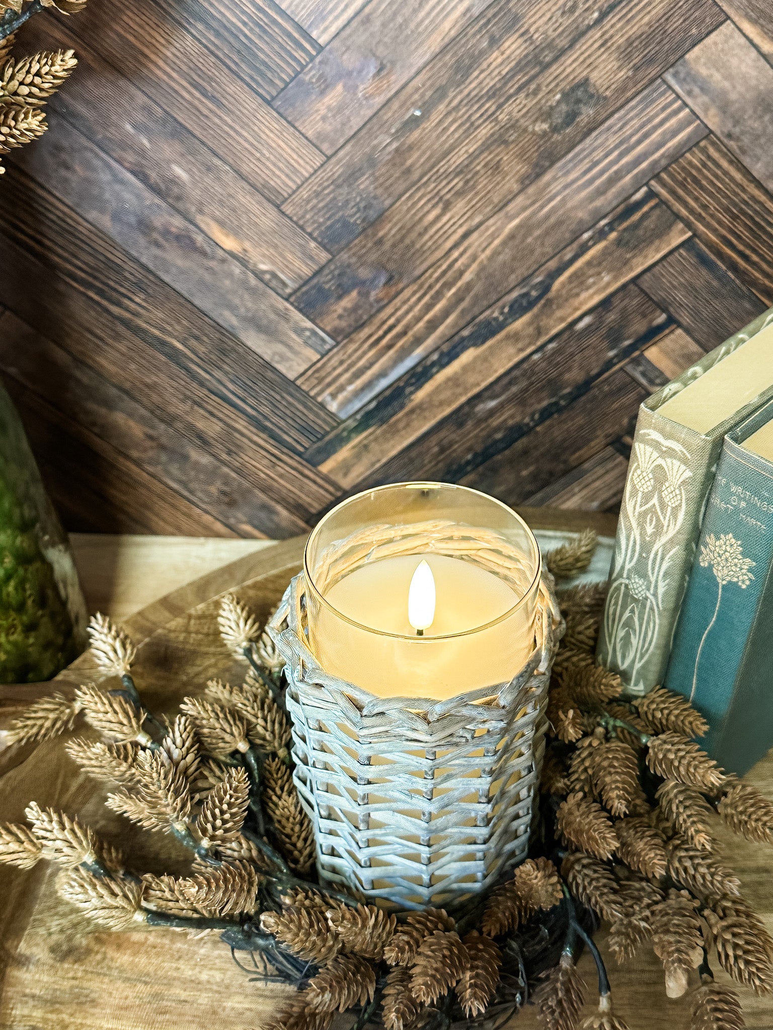 A lit candle in a woven light gray holder sits on a wooden surface surrounded by dried pinecones and branches, with a dark wood herringbone wall and books in the background at the rustic barn in killingworth, CT