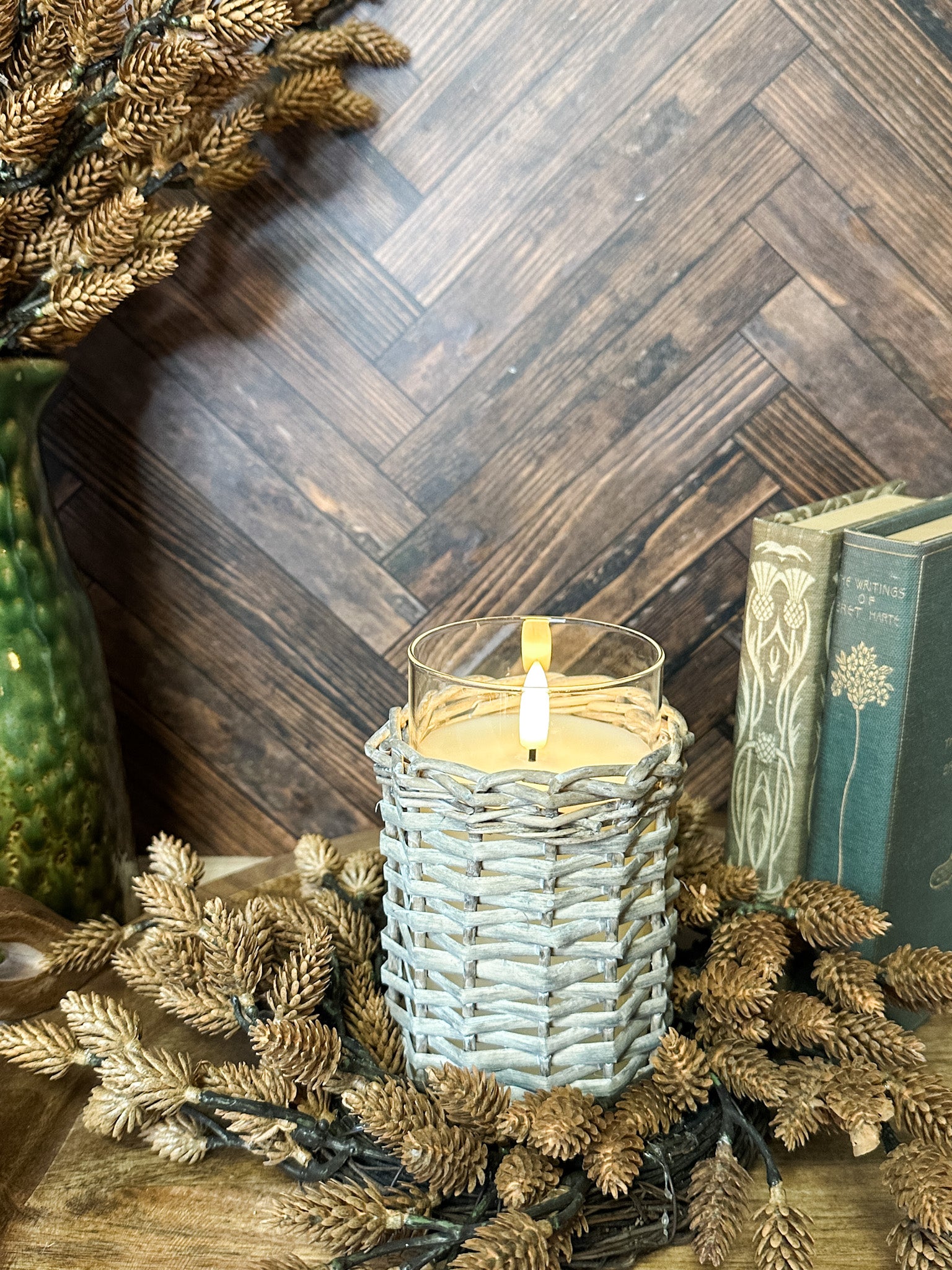 A lit candle in a woven light gray holder sits on a wooden surface surrounded by dried pinecones and branches, with a dark wood herringbone wall and books in the background at the rustic barn in killingworth, CT