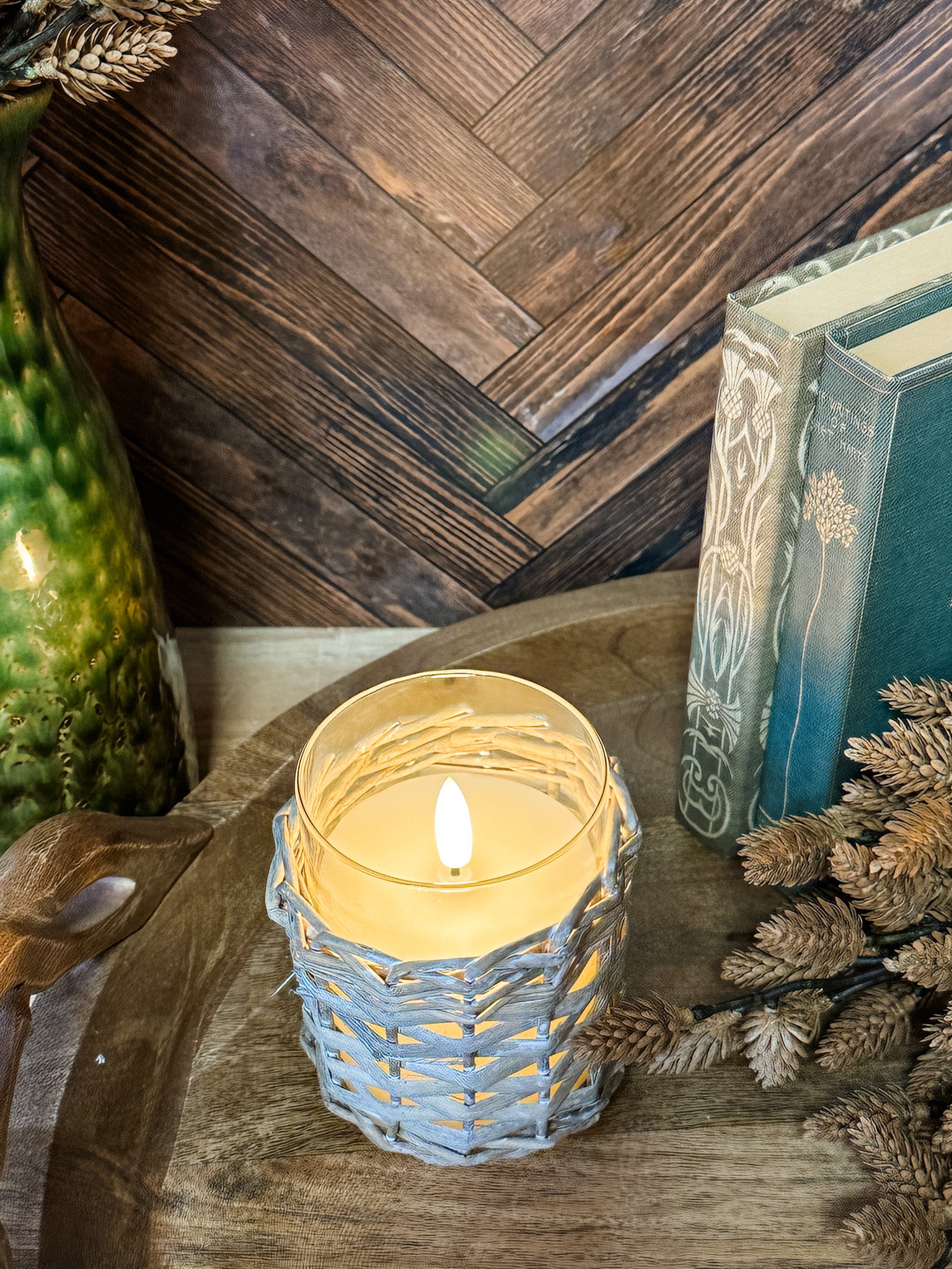 A lit candle in a woven light gray holder sits on a wooden surface surrounded by dried pinecones and branches, with a dark wood herringbone wall and books in the background at the rustic barn in killingworth, CT