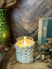 A lit candle in a woven light gray holder sits on a wooden surface surrounded by dried pinecones and branches, with a dark wood herringbone wall and books in the background at the rustic barn in killingworth, CT